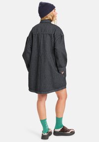 Gestreiftes schwarzes Hemdkleid mit langen Ärmeln, lockerer Passform, Seitentaschen und geschwümmtem Saum, kombiniert mit grünen Socken und gemusterten Schuhen.