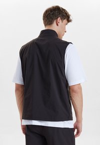 Schwarze ärmellose Weste aus glattem Stoff, mit hohem Kragen, schlichter Gestaltung und lässigem Schnitt, getragen über einem weißen Hemd.