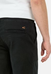 Schwarze Hosen mit glatter Textur, ausgestattet mit einem gestickten Logo und einer Gesäßtasche. Sichtbares Detail des elastischen Bunds.