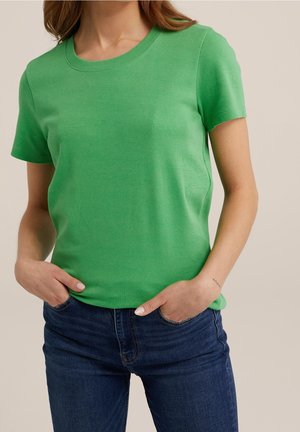 Kobieta w prostym zielonym t-shircie z krótkim rękawem i niebieskich dżinsach z dłońmi w kieszeniach na neutralnym tle.