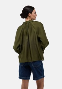 Blouse vert olive avec un détail de pli au dos, manches longues et col. Associée à un short en denim bleu. Texture lisse, coupe ample.