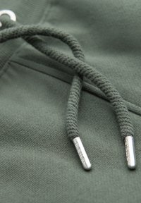 Gros plan sur un tissu de sweat à capuche vert foncé avec des cordons tressés se terminant par des embouts métalliques argentés.