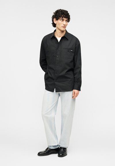 Μαύρο πουκάμισο με κουμπιά, μακριά μανίκια, τσέπη στο στήθος και υφή φινιρίσματος. Φορεμένο με ανοιχτό μπλε τζιν παντελόνι και μαύρα παπούτσια.