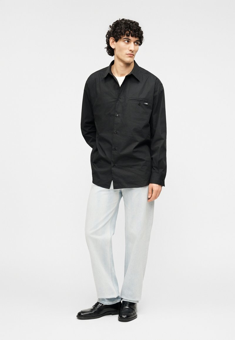 Chemise noire boutonnée avec manches longues, une poche poitrine et une finition texturée. Portée avec un jean bleu clair et des chaussures noires.