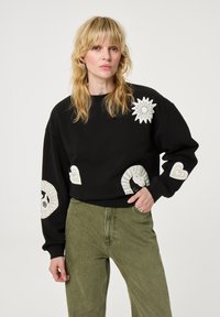 Zwarte sweatshirt met witte grafische accenten, waaronder een zonnestraal en harten, met een ontspannen pasvorm en geribde manchetten, gedragen met olijfgroene broek.