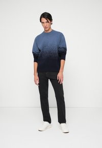 Sweater azul em gradiente com base em azul-marinho escura, tecido de malha leve, punhos e bainha canelados, combinado com jeans pretos e ténis brancos.