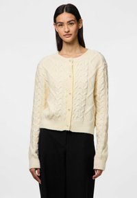 Cardigan crema in maglia a treccia con scollatura rotonda, polsini a coste e chiusura frontale con bottoni decorati da perle. Indossato su pantaloni neri.