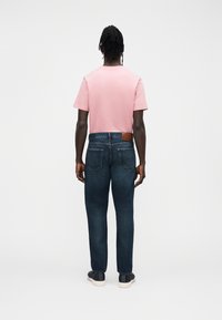 Tagantvaade inimesest, kes kannab helesinist t-särki ja tumedasiniseid teksapükse. Teksadel on vöökohal nahast märk ja kaks tagataskut.