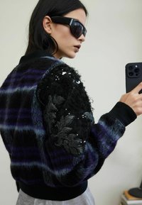 Rozmyta kurtka bomberka w fioletowo-czarną kratę, z teksturowanymi czarnymi rękawami z cekinami i haftem liści. Ściągacze na mankietach i dole.