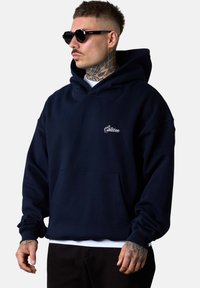 Sweatshirt à capuche bleu marine avec une coupe décontractée, doté d'une poche frontale et d'un logo brodé. Le matériau semble doux avec des poignets côtelés.