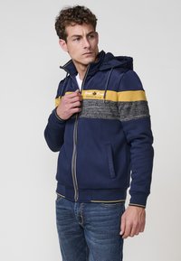 Sudadera con cremallera en color navy, con detalles a rayas amarillas y grises, sección media texturizada, bolsillos laterales y capucha con cordón. Presenta el logo en el pecho.