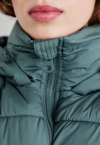Padded jakna u mutnom tirkiznom s visokom ovratnikom i zatvaranjem na patentni zatvarač; ima mekanu teksturu i suptilne detalje šivanja.