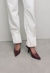 Chaussures à talons hauts en imprimé serpent rouge avec un bout pointu et des côtés ouverts, associées à un pantalon blanc à jambes droites. Texture lisse et design épuré.