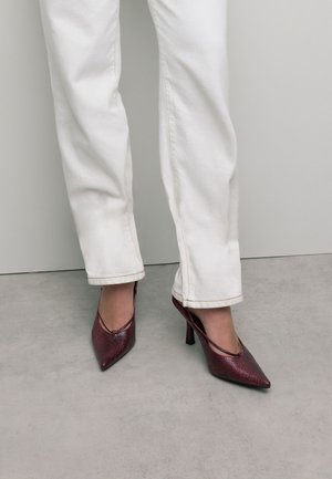 Rote Schlangenleder-High-Heels mit spitzer Zehenform und offenen Seiten, kombiniert mit weißen, gerade geschnittenen Hosen. Glatte Textur und elegantes Design.