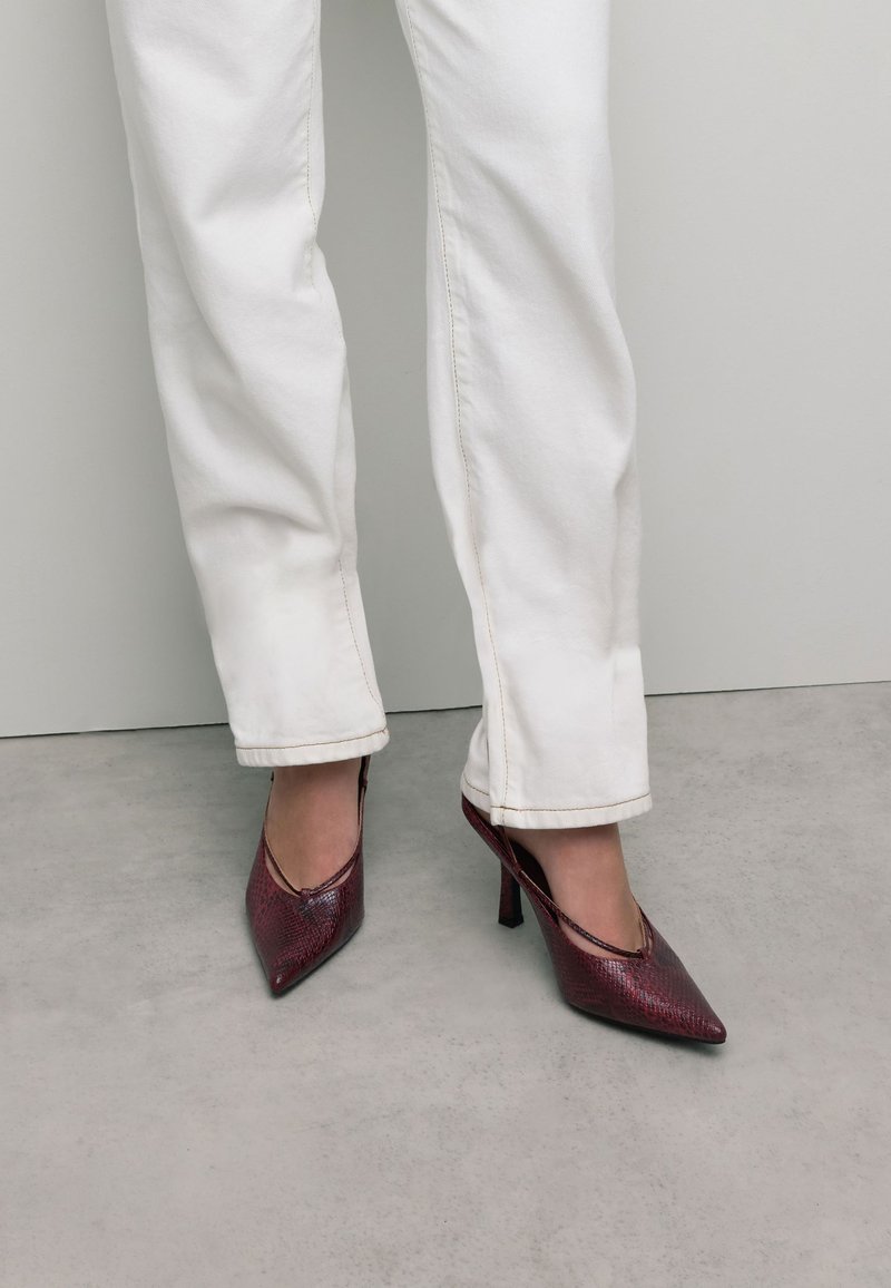 Chaussures à talons hauts en imprimé serpent rouge avec un bout pointu et des côtés ouverts, associées à un pantalon blanc à jambes droites. Texture lisse et design épuré.