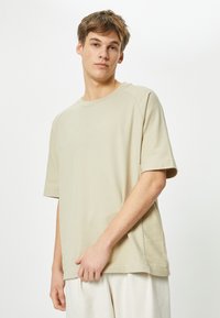 Beige lyhythihainen t-paita, jossa on pyöreä pääntie ja rento malli, valmistettu pehmeästä kankaasta. Ominaisuuksiin kuuluu hienovarainen tekstuuri ja sivusaumat.