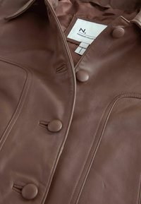 Veste en cuir marron avec des boutons recouverts de tissu et une étiquette blanche portant l'inscription "Premium Collection" à l'intérieur du col.