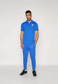Mann står iført en blå Nike-polo og matchende joggebukser med Atlético Madrid-logoer og svarte joggesko mot en enkel lys bakgrunn.