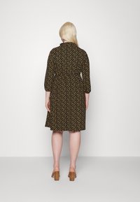 Vestido preto com um pequeno padrão floral amarelo, mangas três quartos, colarinho e comprimento médio. Usado com sapatos de salto em tom bege.
