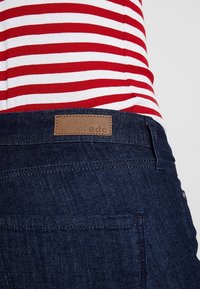 Närbild av en person som bär mörkblå denimbyxor med en brun "edc"-etikett och en röd-vit randig skjorta instoppad.