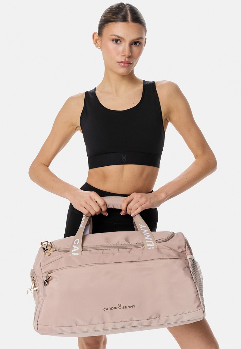 Kobieta w czarnym sportowym biustonoszu trzyma beżową torbę na siłownię Cardio Bunny za uchwyty na jednolitym jasnym tle.