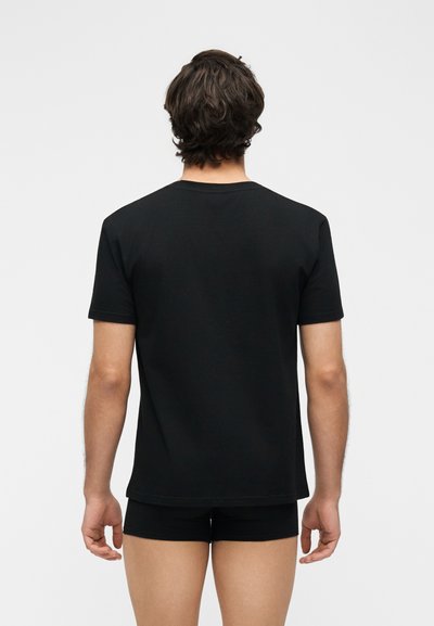 Άνδρας με σκούρα μαλλιά, φορώντας ένα απλό μαύρο κοντομάνικο μπλουζάκι και μαύρα σορτς, στέκεται με την πλάτη προς τα έξω σε ανοιχτό φόντο.