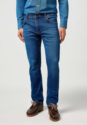 Mężczyzna ubrany w niebieskie jeansy z denimu, niebieską dżinsową koszulę oraz brązowe skórzane buty sznurowane, stojący na jednolitym tle.