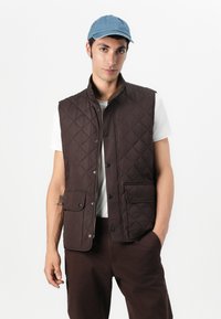 Καφέ επενδυμένο γιλέκο με ψηλό γιακά, μπροστινά κουμπιά και δύο μπροστινές τσέπες, συνδυασμένο με λευκό πουκάμισο και μπλε τζην καπέλο.