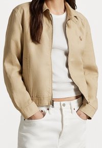 Béžová, lehká bunda s límcem, zapínáním na zip a elastickými manžetami, v kombinaci s bílým žebrovaným topem a bílými džíny.