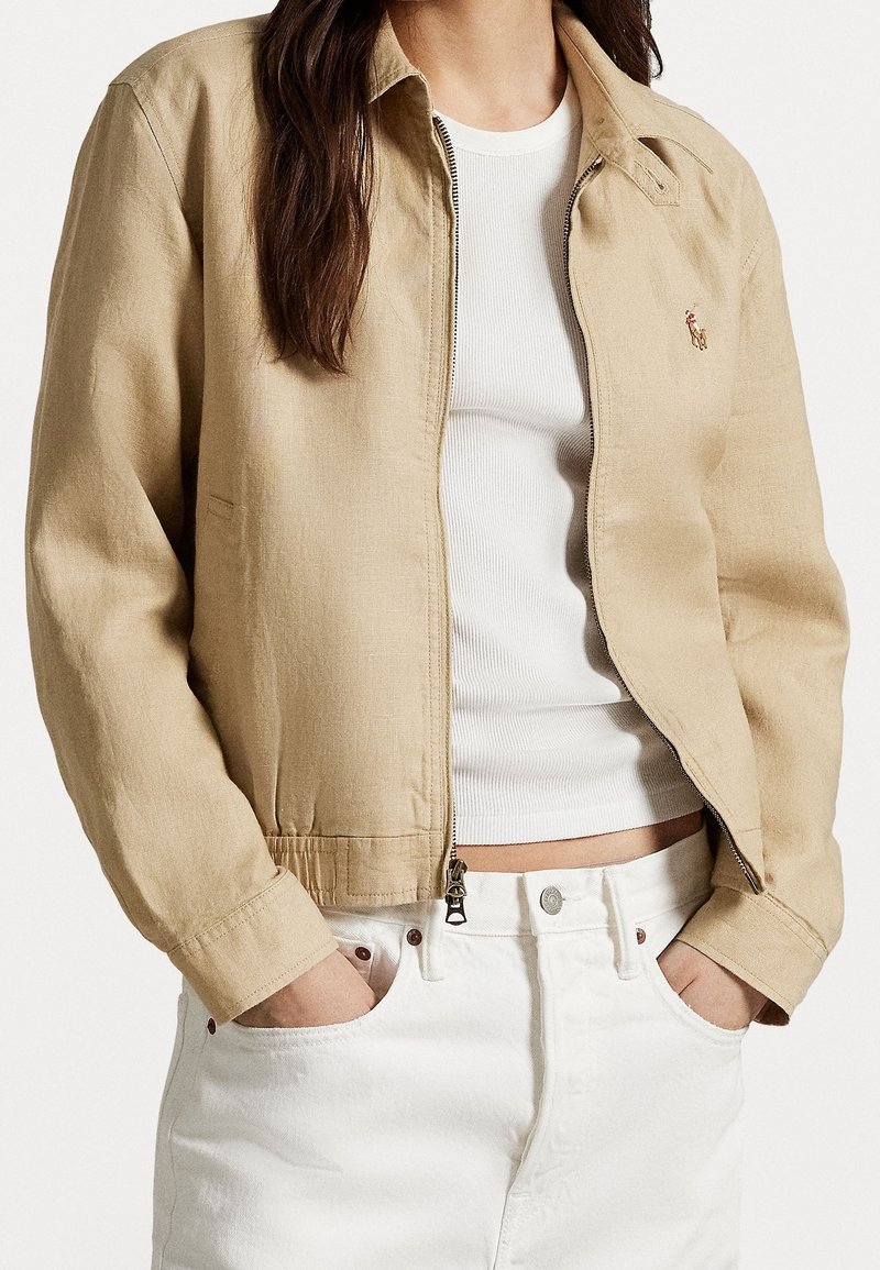 Béžová, lehká bunda s límcem, zapínáním na zip a elastickými manžetami, v kombinaci s bílým žebrovaným topem a bílými džíny.