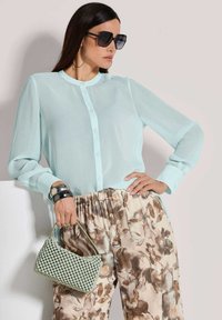 Himmelblaue, durchsichtige Bluse mit Knopfleiste und langen Ärmeln; kombiniert mit floral gemusterten, elastischen Hose. Grüner, strukturierten Handtasche und übergroßen Sonnenbrille.