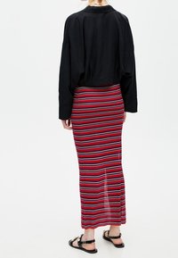 Černá oversized košile s dlouhými rukávy v kombinaci s maxi sukní s červenými a bílými pruhy. Sukně má boční rozparek a přiléhavou siluetu.