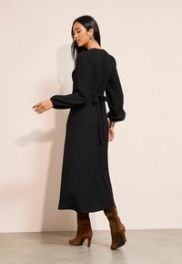Čierne midi šaty s textúrou, puffovými rukávmi a viazaným pásom. Model má na sebe hnedé semišové čižmy na podpätku. Minimalistický dizajn na neutrálne pozadí.