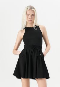 Svart kjole med tettsittende ribbestrikket liv, smocket midje, flagrende skjørt og lommer i sidene. Glatt tekstur, avslappet design og justerbar snor.