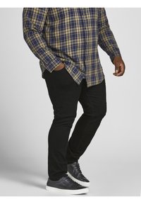 Ruudullinen paita tummansinisenä, vihreänä ja keltaisena; kapeat mustat housut; mustat tennarit valkoisilla pohjilla; kädet taskuissa; sivuprofiili.