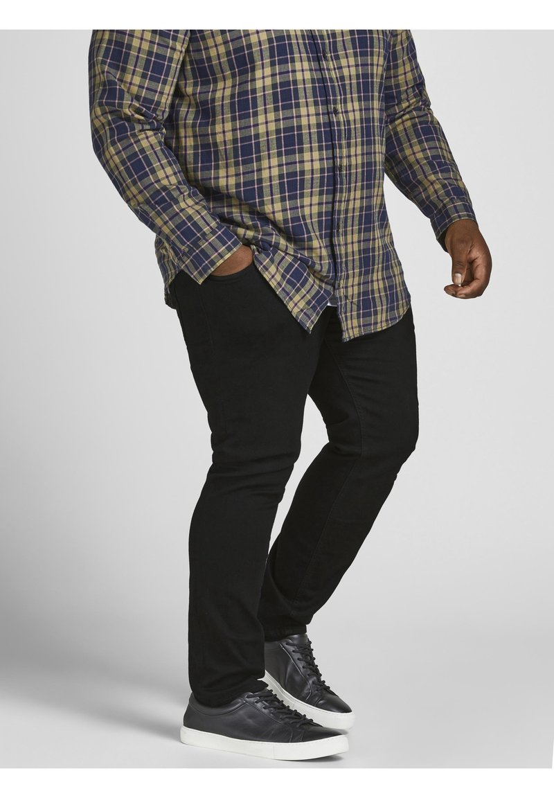 Ruudullinen paita tummansinisenä, vihreänä ja keltaisena; kapeat mustat housut; mustat tennarit valkoisilla pohjilla; kädet taskuissa; sivuprofiili.