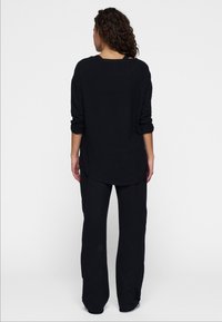 Schwarzes Langarmoberteil mit abgerundetem Saum und hochgekrempelten Ärmeln, kombiniert mit schwarzen, locker sitzenden Hosen. Glatte Textur, minimalistisches Design.