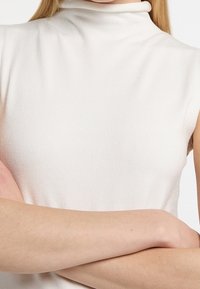 Ärmelloses Rollkragen-Top in hellem Creme, mit geripptem Muster und figurnaher Passform. Glattes Material und minimale Nahtdetails.