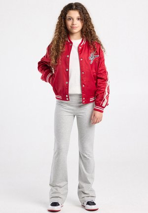 Jong meisje met lang krullend haar, gekleed in een rode varsityjas, wit shirt, lichtgrijze broek en zwart-witte sneakers, staand tegen een witte achtergrond.