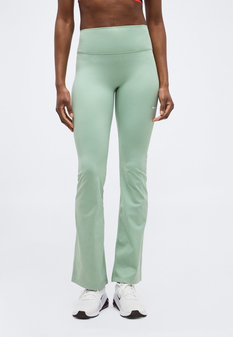 Femme portant un pantalon de sport évasé vert clair et des baskets blanches, debout les mains détendues le long du corps sur un fond uni.