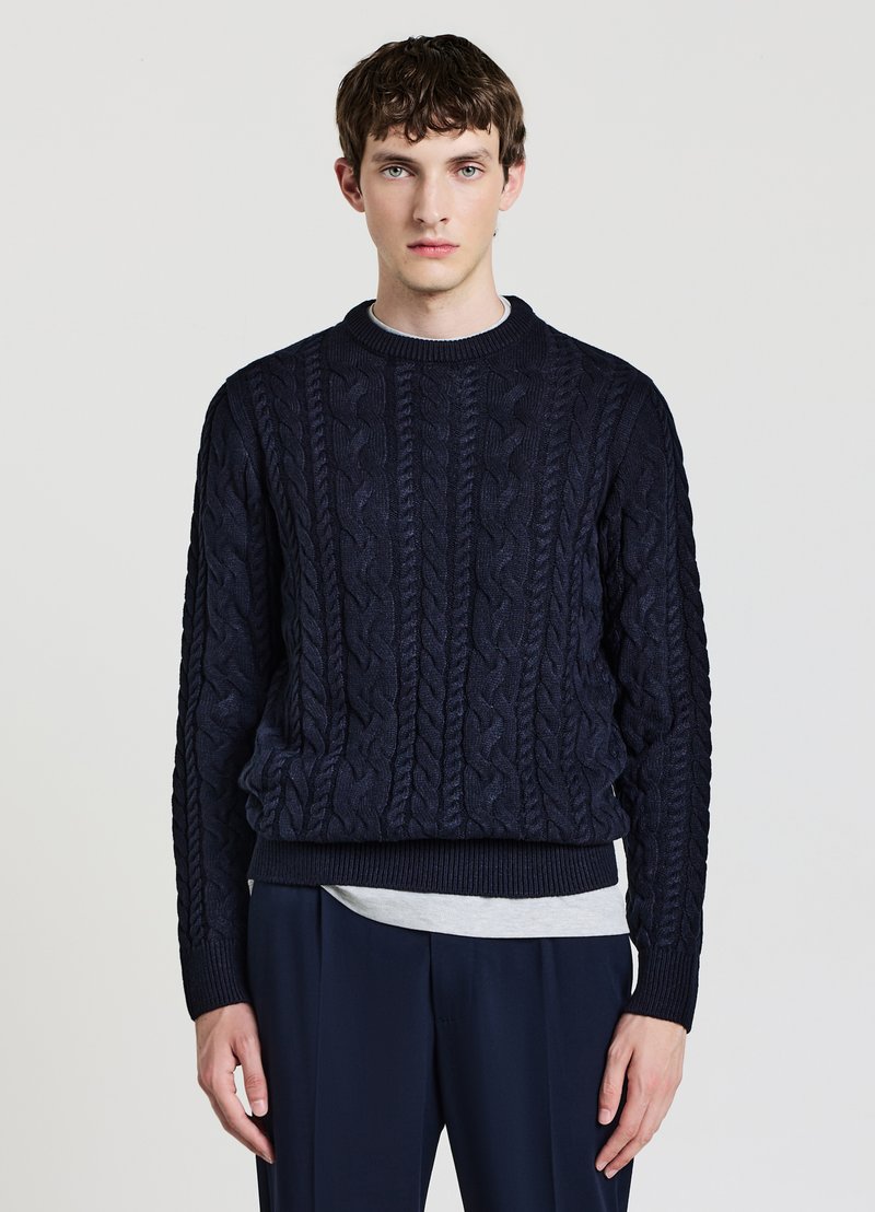 Marineblauer Pullover mit Zopfmuster, rundem Ausschnitt, strukturiertem Muster, gerippten Bündchen und Saum. Trägt darunter ein hellgraues Hemd, kombiniert mit dunklen Hosen.