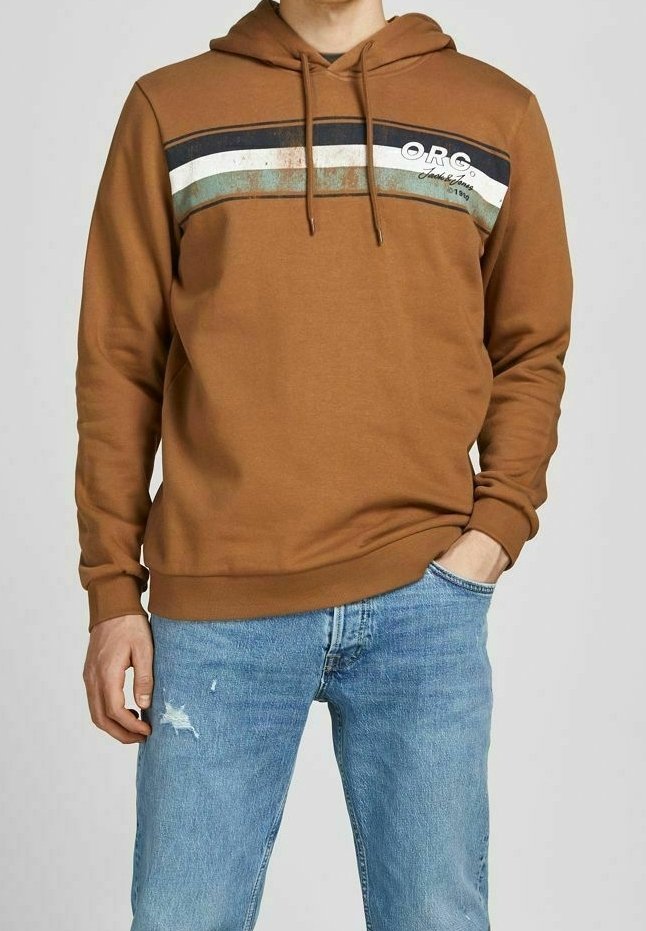 Homme en sweat à capuche marron avec des rayures sur la poitrine et un jean bleu, debout une main dans la poche devant un fond uni.