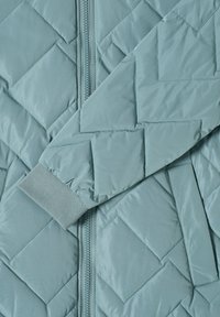 Manche et devant d'une veste matelassée bleu clair avec piqûres en losange, poignet côtelé, fermeture éclair avant et poche latérale.