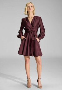 Robe portefeuille bourgogne en satin doux avec de longues manches bouffantes, une taille à nouer, une jupe évasée et des poches latérales. Portée avec des escarpins à brides.