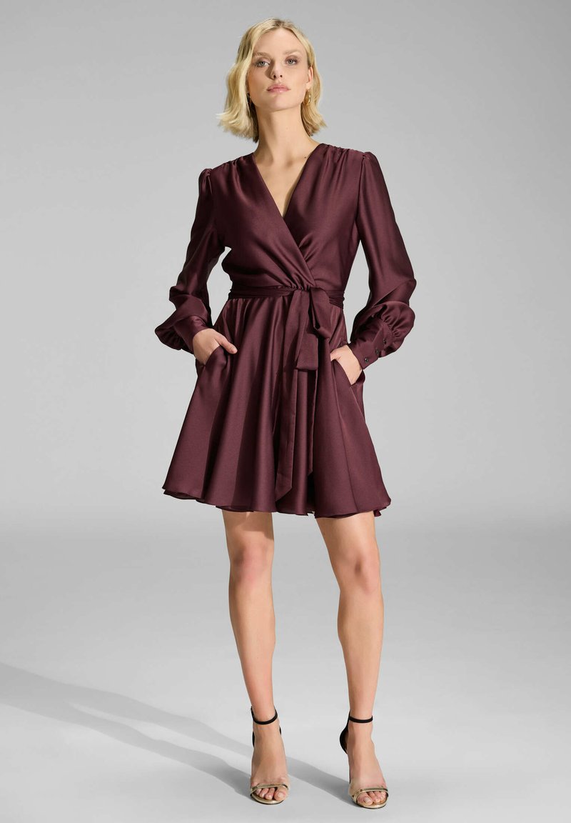 Robe portefeuille bourgogne en satin doux avec de longues manches bouffantes, une taille à nouer, une jupe évasée et des poches latérales. Portée avec des escarpins à brides.
