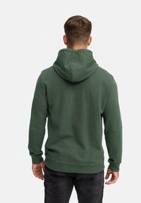 Grøn hættetrøje med struktureret overflade, der har en central søm på ryggen og ribbede manchetter, båret med mørke jeans.