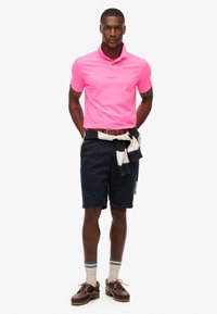 Tenue masculine comprenant un polo rose vif, un short bleu marine, un pull beige noué à la taille, des chaussettes beiges et des chaussures marron.