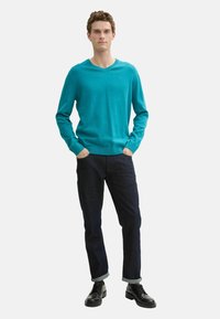 Pulover turcoaz cu guler în V, din tricot fin, asortat cu blugi negri din denim, cu manșete rulate și pantofi negri cu șireturi. Design simplu și ajustat.