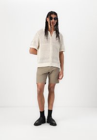 Camicia corta a maniche corte in crochet di colore beige con chiusura a bottoni, abbinata a pantaloni corti color khaki. Scarpe nere e occhiali da sole rotondi completano l'outfit.