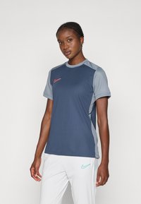 Nainen, jolla on yllään tummansininen ja harmaa Nike-urheilupaita ja valkoiset housut, joissa on turkoosi Nike-logo, seisoo yksinkertaista taustaa vasten.